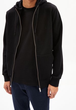 Homme portant un sweat à capuche zippé noir avec des cordons sur un t-shirt noir et un pantalon bleu foncé, debout devant un fond blanc.