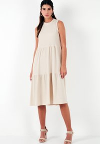 Beige sleeveless dress with a high neckline, tiered skirt, and textured fabric. Accented with neutral heeled sandals.