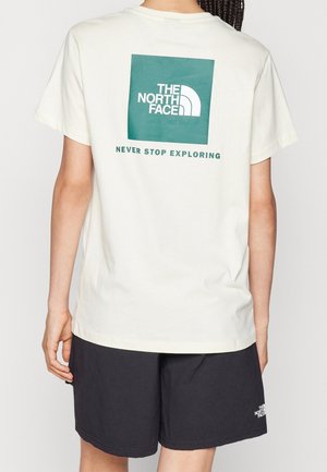 Person trägt ein weißes T-Shirt mit grünem "The North Face"-Logo und dem Schriftzug "Never Stop Exploring" auf dem Rücken, kombiniert mit schwarzen Shorts.