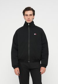 Black zip-up bomber jacket with a high collar, ribbed cuffs, and hem, featuring a small red, white, and blue logo on the left chest.