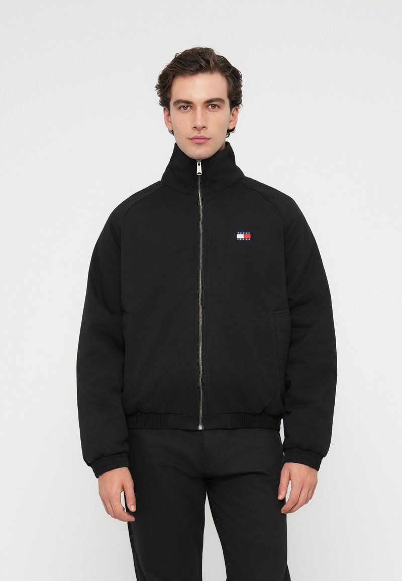 Black zip-up bomber jacket with a high collar, ribbed cuffs, and hem, featuring a small red, white, and blue logo on the left chest.