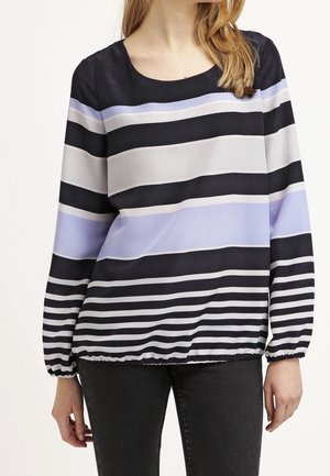 Vrouw draagt een blouse met lange mouwen en brede horizontale strepen in zwart, wit en lichtpaars, gecombineerd met een zwarte broek.