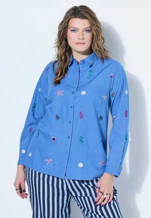 Kvinde med bølget hår iført en blå skjorte med knapper og broderede havdyr samt blå og hvide stribede bukser.