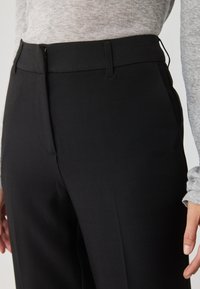 Calças pretas sob medida com uma textura suave, apresentando um fecho frontal e passantes para cinto. Combinadas com uma camisa de manga longa cinza claro.