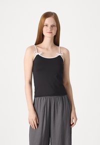 Svart tanktopp med hvit kant og justerbare stropper knyttet i sløyfer. Myk stoff; paret med grå vide bukser. Enkelt og allsidig design.