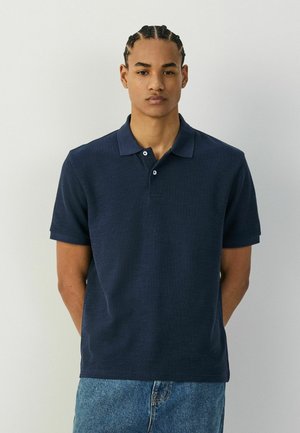 Junger Mann mit geflochtenen Haaren, der ein dunkelblaues Polohemd und helle blaue Jeans trägt und mit den Händen hinter dem Rücken vor einem einfarbigen Hintergrund steht.