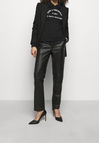 Mujer vestida con pantalones de cuero negros, tacones altos negros, blazer negro y sudadera con capucha negra con texto blanco, de pie contra un fondo liso.