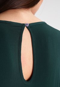 Rückenansatz eines dunkelgrünen, strukturierten Blusentops mit einem einzelnen lila Knopfverschluss und einer ovalen Schlüssellochöffnung, die Haut zeigt.