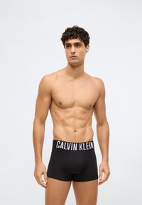 Kailu bez krekla vīrietis ar īsiem cirtaini mati, ģērbies melnās Calvin Klein bokseršortos, stāv uz vienkārša gaiši pelēka fona.