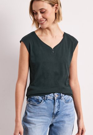 Mujer sonriente que lleva una blusa sin mangas de cuello en V de color verde oscuro con dos botones y jeans azul claro, de pie frente a un fondo liso.