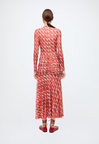 Robe rouge à motifs avec manches longues, présentant une taille froncée et une jupe fluide. Le tissu est léger et semi-transparent. Portée avec des ballerines rouges.