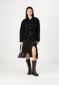 Schwarze Jeansjacke mit Knopfverschlüssen, über einem schwarzen asymmetrischen Rock getragen. Braune knielange Stiefel mit Schnallen, dazu eine schwarze Handtasche.
