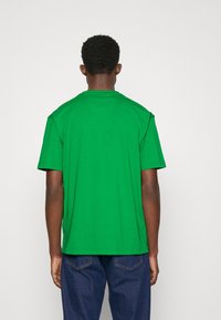 Zelené bavlněné tričko s krátkým rukávem a kulatým výstřihem. Má jednoduchý design a rovný lem, v kombinaci s modrými džínami.