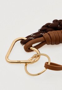 Καφέ πλεκτός σκοινί με δερμάτινες λεπτομέρειες, χρυσό τριγωνικό κλιπ και κυκλικό δαχτυλίδι. Λείος υφή με αντίθετες λευκές ραφές.