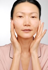 Una donna in un accappatoio rosa chiaro applica una crema sul viso, tenendosi le guance con entrambe le mani. Ha la pelle liscia e un trucco naturale.
