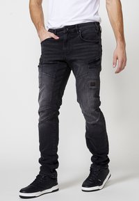 Črne jeans cargo hlače z ožjim krojem, sprednjimi žepi in dodatnimi stranskimi žepi. Teksture tkanine z diskretnim zbledelostjo in detajli šivanja.