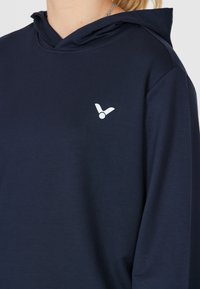 Mörkblå hoodie i mjukt material, med en kenguruficka och en liten vit logotyp på vänster bröstområde. Inkluderar en dragsko i huvan.