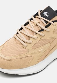 Beige sportcipő, textúrázott hálós felsőrésszel, velúr részletekkel, fekete fűzőkkel és fehér talppal, amely oldalán dizájnelemek találhatók.