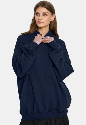 Młoda kobieta z długimi brązowymi włosami, ubrana w luźną, granatową bluzę z kapturem, trzymająca obie ręce na kołnierzu, spoglądająca lekko w prawo.