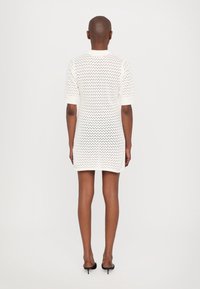 Robe blanche courte avec un design perforé, dotée de manches courtes et d'une silhouette ajustée. Associée à des chaussures à talons hauts noires.