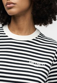 Άτομο που φοράει μαύρο και άσπρο ριγέ πουκάμισο Carhartt WIP, με ορατό λαιμό, γραμμή σαγονιού και σγουρά μαλλιά πάνω σε λευκό φόντο.