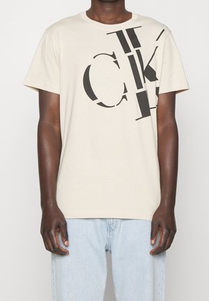 T-shirt beige à manches courtes en coton, avec de grandes lettres graphiques noires sur le devant. Jean en denim bleu clair porté.