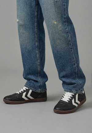 Gambe con jeans blu sbiaditi e sneakers nere con strisce bianche a forma di chevron e suole marroni su sfondo grigio.