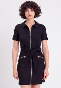 Vestido negro de manga corta con cremallera frontal, cuello, cinturon en la cintura y dos bolsillos con cremallera. Hecho de una tela texturizada.