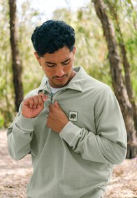 Suéter verde claro hecho de una tela suave, con un cuello alto, cremallera frontal, puños acanalados y un pequeño logo de elefante en el pecho.