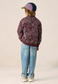 Sweatshirt bordeaux avec des illustrations de mains blanches et du texte. Jean large bleu clair, avec des ourlets roulés, et des baskets blanches avec des accents gris. Casquette violette.