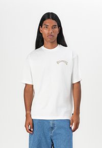 Hvid bomulds t-shirt med rund hals og korte ærmer, der har et farverigt broderet logo på brystet. Båret med blå denimshorts.