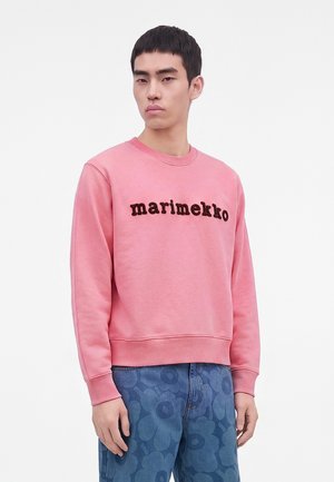 Млад мъж, облечен в розова суитшърт Marimekko и сини дънки с флорални мотиви, стои пред обикновен светъл фон.