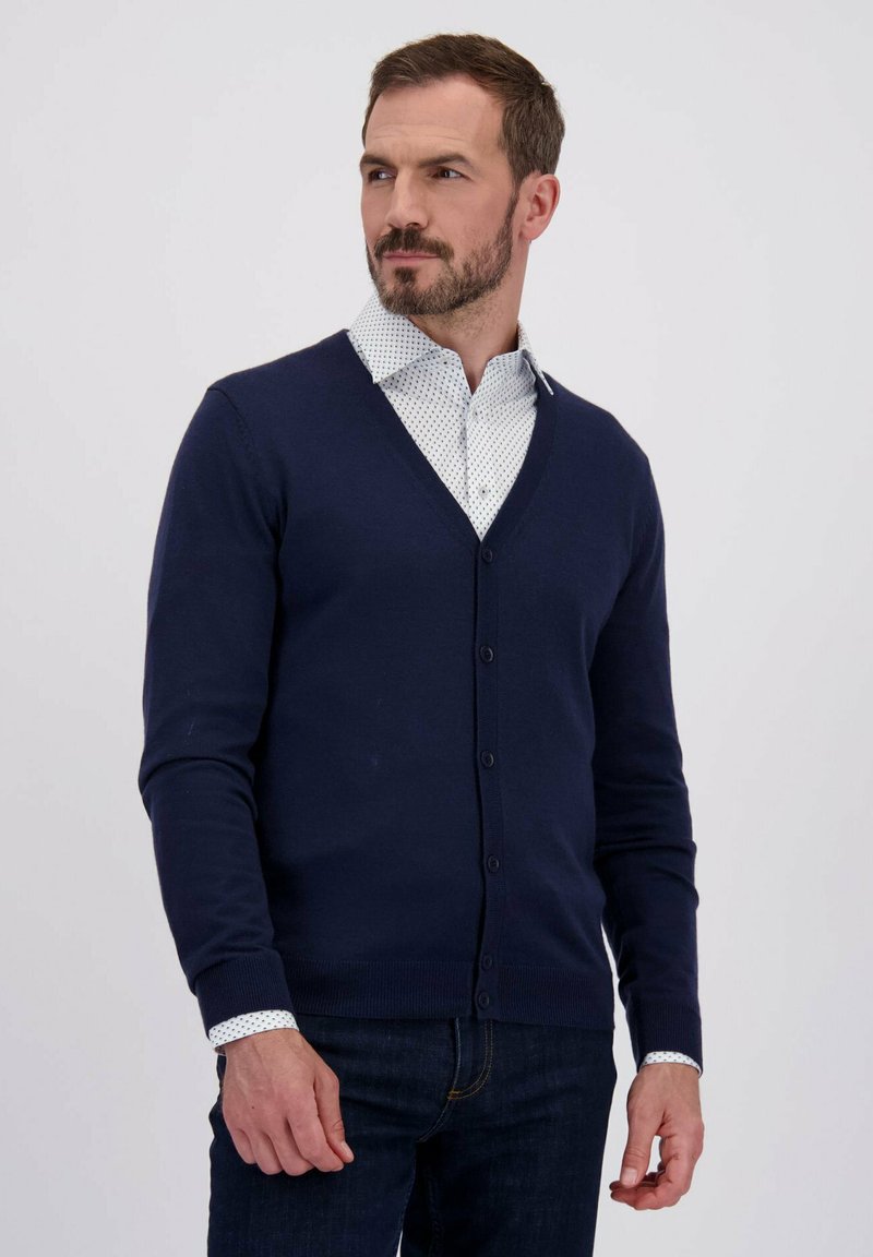 Man in een navyblauwe gebuttonde cardigan over een witte gepatternde shirt, poserend voor een effen lichte achtergrond.