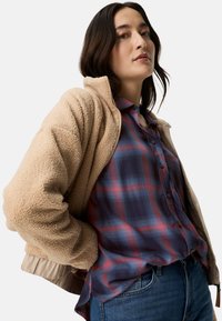 Beige textured fleece jacket with a zipper and ribbed cuffs, layered over a blue and red plaid shirt, paired with denim jeans.