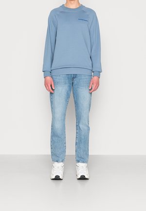 Sweatshirt bleu clair à col rond avec des poignets et un ourlet côtelés, arborant un petit logo sur la poitrine. Associé à un jean droit bleu clair et des baskets blanches.