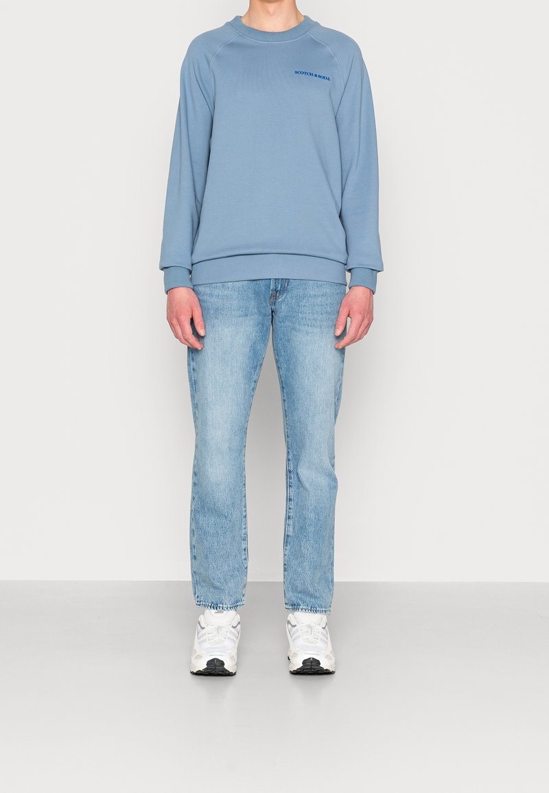 Světle modrý crewneck sweatshirt s žebrovanými manžetami a lemem, na hrudi s malým logem. V kombinaci se světle modrými džíny rovného střihu a bílými teniskami.
