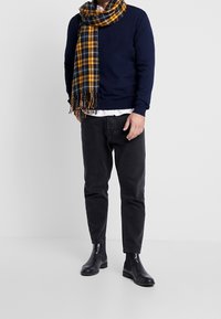 Marinblå tröja, rutigt gult och svart halsduk, svarta jeans och svarta ankelskor visade på en modell mot en enkel bakgrund.