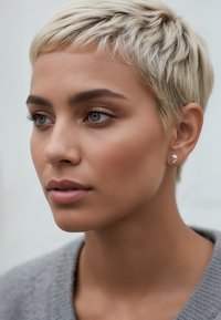 Nahaufnahme eines Gesichts mit kurzen blonden Haaren. Es hat einen silbernen Totenkopf-Studiowellen. Der Hautton ist warm, mit dezentem Make-up und einem grauen Pullover.