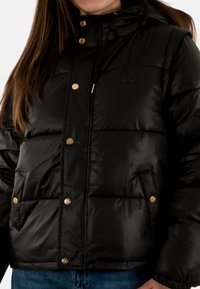 Giacca a vento nera con cappuccio, caratterizzata da una texture lucida, hardware dorato, tasche frontali e un logo in rilievo sul petto.