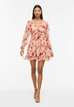 Ženska, oblečena v roza cvetlico dolgo rokavno mini obleko z volančki in zlatimi sandali z visokimi petami, stoji ob belem ozadju.