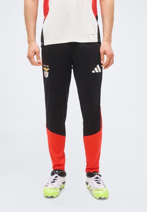 Personne portant un pantalon de sport noir et rouge avec les logos Adidas et Benfica, des crampons blancs avec des semelles vert fluo et un top de sport blanc.