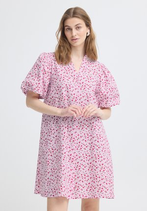 Kobieta w różowej sukience z drobnym czerwonym kwiatowym wzorem i bufkami na krótkich rękawach, stojąca na jednolitym jasnym tle.