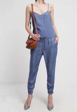 Donna che indossa una tuta senza maniche in denim blu con cintura a coulisse in vita, borsa marrone a tracolla e décolleté con motivo a pelle di serpente.