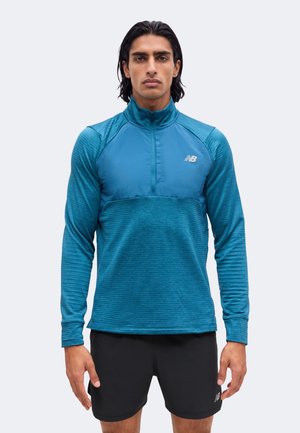 Jeune homme aux cheveux mi-longs foncés portant un haut à manches longues bleu à demi-fermeture éclair et un short noir sur un fond clair.