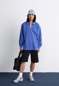 Blue cotton pullover with a zip neck, paired with black pinstriped shorts. Accessories include a light blue bucket hat and a black tote bag.