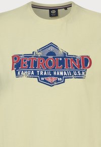 Světle žluté bavlněné tričko s modrým a červeným opotřebovaným potiskem obsahujícím text "PETROL IND" a "KAHUA TRAIL HAWAII U.S.A.".