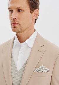 Beżowa marynarka z białą koszulą i kamizelką w kratkę houndstooth. Poszetka ma wzór w kropki w odcieniach bieli i szarości.