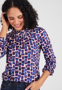 Blusa a maniche lunghe con collo alto arricciato, caratterizzata da un motivo geometrico in rosso, blu scuro e blu su uno sfondo crema.
