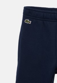 Tumši zili treniņbikses ar elastīgu jostu, kurām pie gurniem ir izšūta maza zaļa krokodila logo.