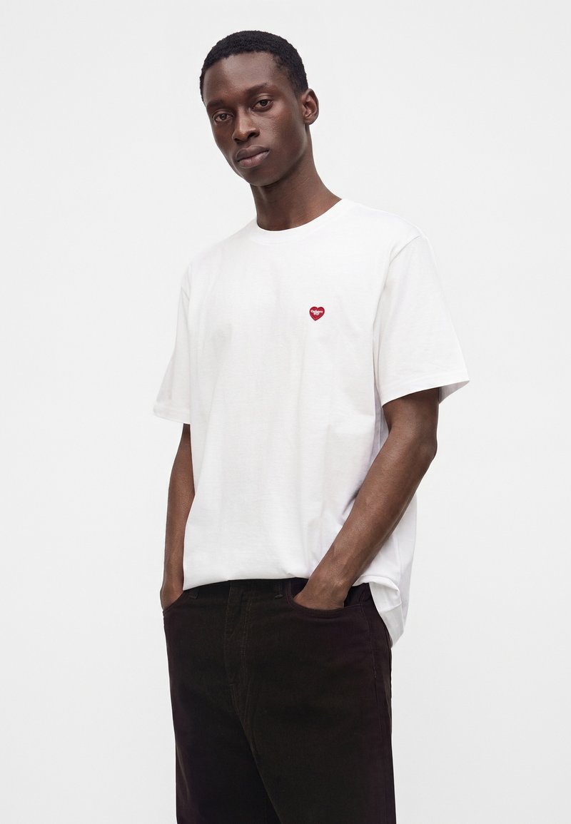 Junger Mann trägt ein weißes T-Shirt mit kleinem rotem Herz-Emblem auf der linken Brustseite und schwarze Hose, steht mit den Händen in den Taschen.